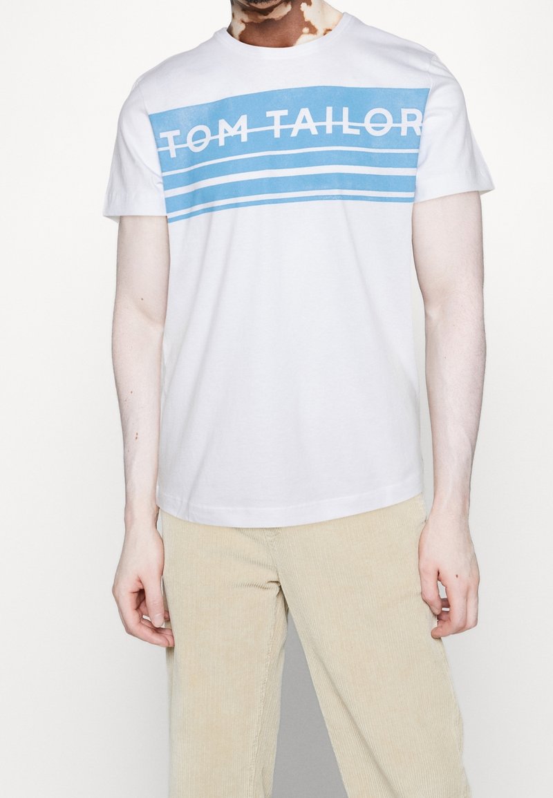 T-shirt en coton blanc avec des rayures horizontales bleues et le texte "TOM TAILOR" en blanc. Manches courtes et col rond.