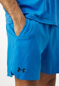 Blaue Sportshorts aus leichtem Stoff mit glatter Textur. Mit Seitentaschen und einem schwarzen Logo auf der unteren linken Seite.