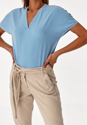 Kobieta w jasnoniebieskim topie z krótkim rękawem i dekoltem w serek, wsadzonym w beżowe spodnie z wysokim stanem i materiałowym paskiem zawiązanym na supeł, z rękami opartymi na biodrach.