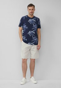 Marineblauwe T-shirt met witte palmboomprint, gecombineerd met lichtbeige shorts. Witte sneakers maken de look af. Eenvoudige, casual outfit.