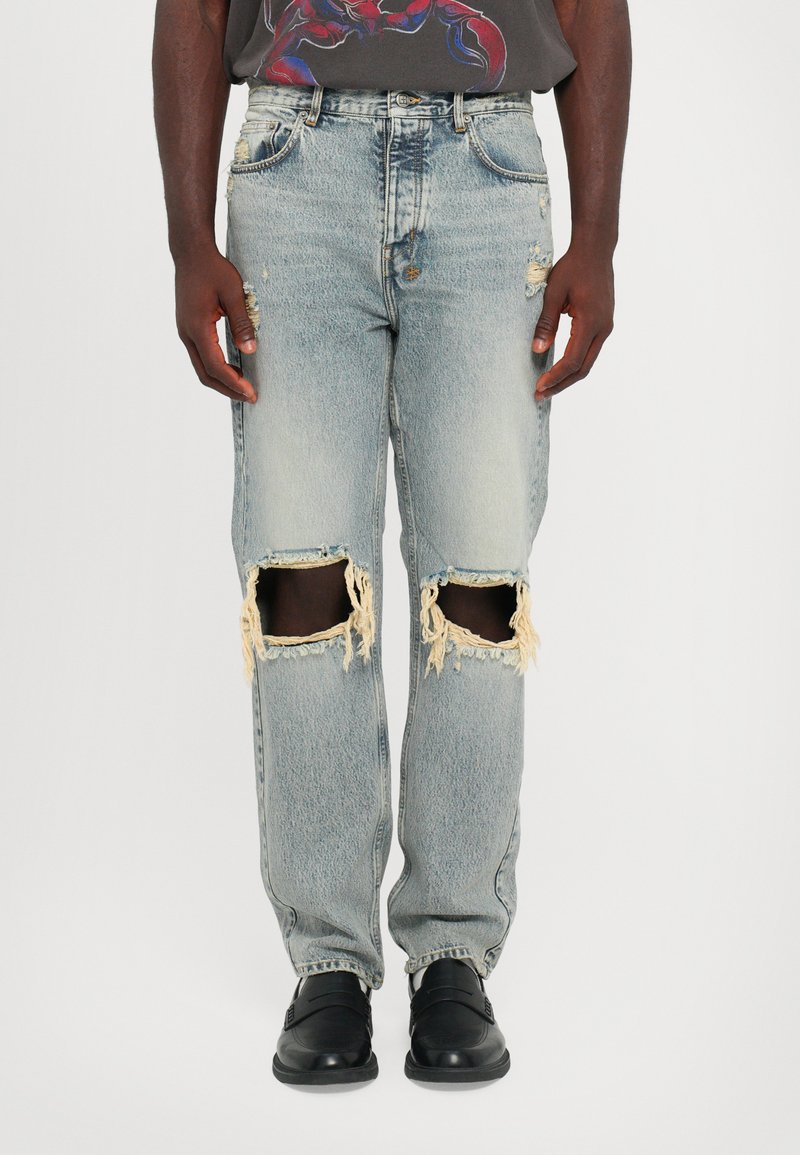 Jeans din denim decolorat, de culoare albastră deschisă, cu mari rupturi la genunchi, margini desfăcute și o croială lejeră. Combinat cu pantofi negri slip-on.