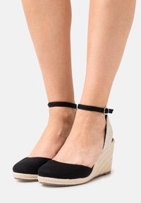 Scarpe con tacco a cuneo in tessuto nero con tacco in beige intrecciato e cinturino alla caviglia con dettaglio in fibbia argentata.