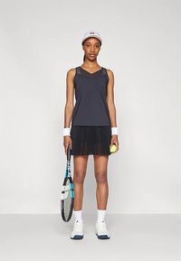 Μαύρη αμάνικη μπλούζα με V-λαιμόκοψη, πλισέ μαύρη φούστα, λευκά βραχιολάκια και καπέλο. Κρατάει ένα ρακέτα τένις και μια μπάλα τένις.