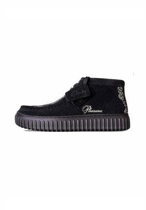 Black suede ankle boots with a ribbed rubber sole. Features stitching details and embroidered "Clarks" and "Pleasures" logos.