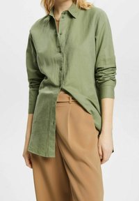 Camicia verde oliva con bottoni, realizzata in materiale morbido, con maniche lunghe e vestibilità rilassata, abbinata a pantaloni beige.
