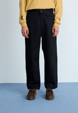 Pantalones de pierna ancha negros con cintura alta, que cuentan con un cierre de botón en el frente y bolsillos laterales, combinados con zapatos mocasines marrones.