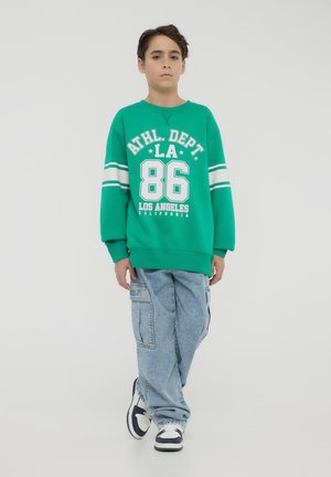 Felpa verde con lettering e strisce bianche, abbinata a jeans cargo azzurri chiari e sneakers nere e bianche. Sfondo semplice.