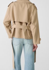 Béžový trenčkot s přizpůsobeným střihem, dvouřadým zapínáním a uvázaným páskem v pase. Obsahuje knoflíkové detaily a dlouhé rukávy.