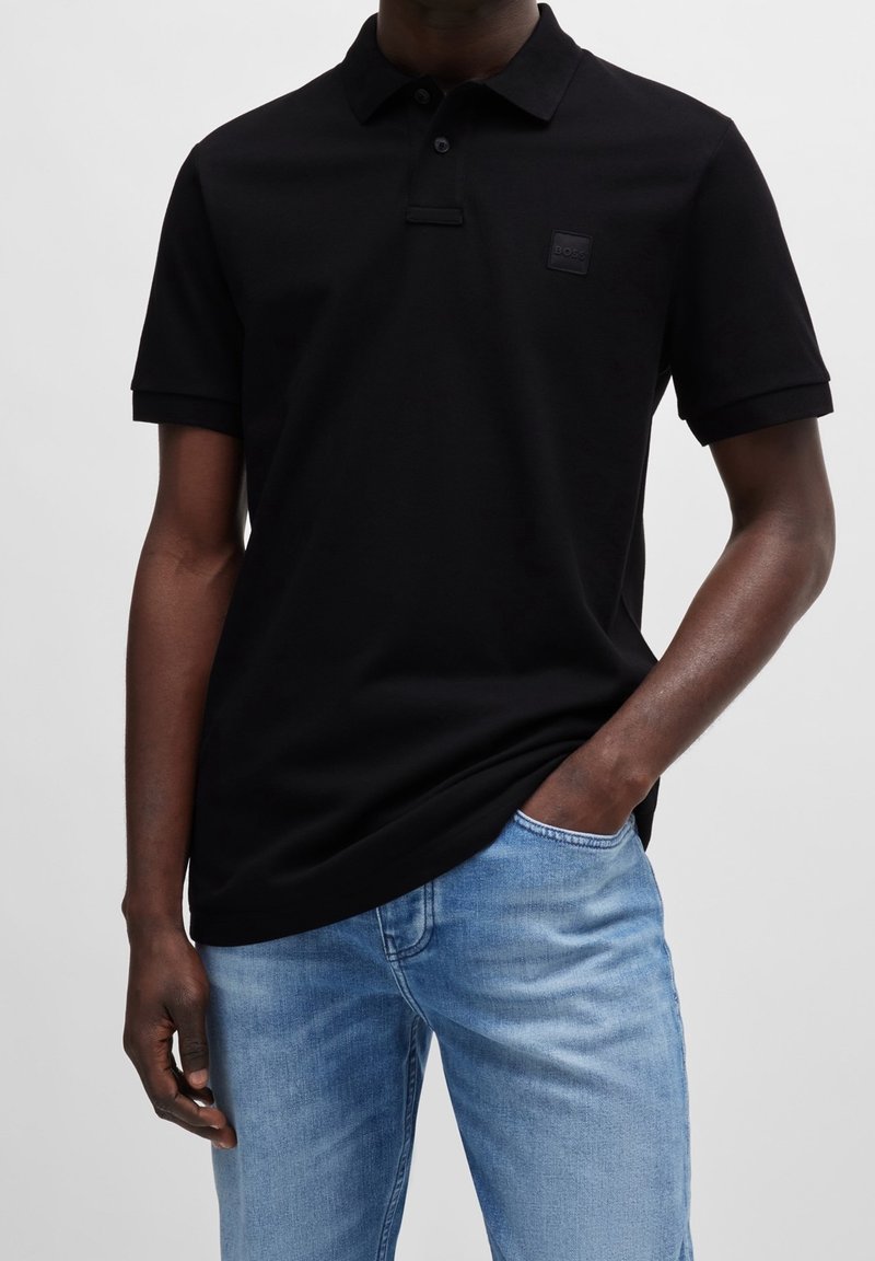 Homme portant un polo noir à manches courtes avec un petit logo sur la poitrine et un jean en denim bleu clair, une main dans la poche.