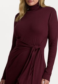 Abito bordeaux a maniche lunghe con collo alto. Presenta un davanti drappeggiato fissato con un nodo, realizzato in tessuto morbido ed elastico.
