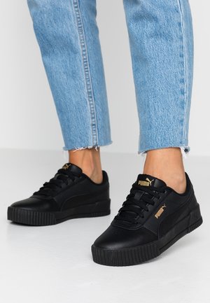 Person trägt schwarze Puma-Sneaker mit goldenen Logos und hellblauen cropped Jeans, steht auf einem weißen Boden.