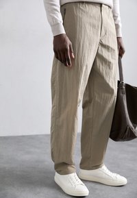 Beige, textured trousers with a relaxed fit, featuring zippered pocket detail and white sneakers. Model holds a dark brown bag.