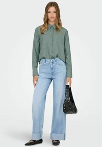 Camisa verde de botones, vaqueros azul claro de pierna ancha con puños doblados, zapatos planos negros y bolso de hombro negro con tachuelas plateadas.