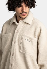 Kurtka koszulowa z kremowego polaru o teksturowanej powierzchni, z dwiema kieszeniami na piersi i białymi guzikami. Posiada haftowane logo na kieszeni na piersi.