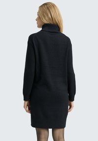 Vrouw met blond haar die een zwart lange mouw turtleneck sweater jurk draagt, met de rug naar de camera op een eenvoudige lichte grijze achtergrond.
