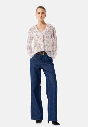 Donna in piedi che indossa una camicetta rosa a pois con volant, jeans blu a gamba larga, cintura nera e tacchi neri a punta.