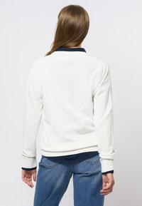 Hvit strikket genser med ribbestrikket nederkant og mansjetter, med en mørk marineblå krage. Brukt sammen med blå denimjeans. Bakfra, glatt tekstur.