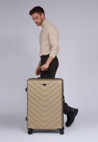 Gouden harde koffer met chevronpatroon, zwarte handgreep en wielen, vastgehouden door een persoon in een beige trui en donkere broek.