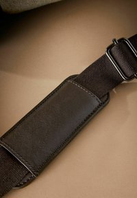 Black adjustable strap with leather accent, featuring smooth texture and contrasting stitching, set against a beige background.