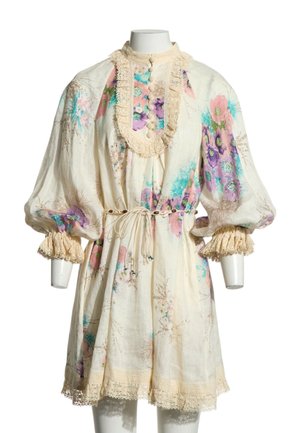 Vestido floral crema con mangas abullonadas, ribete de encaje, cierre con botones en la parte delantera y cinturón de atar en la cintura sobre un maniquí sin cabeza con fondo blanco.