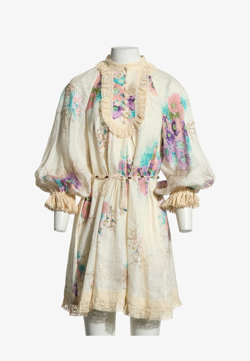 Robe fleurie crème avec manches bouffantes, bordure en dentelle, fermeture boutonnée devant et ceinture nouée à la taille sur un mannequin sans tête sur fond blanc.
