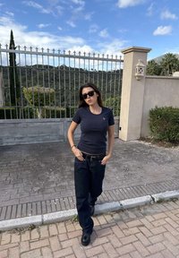 Mladá žena v čiernych slnečných okuliaroch, tmavomodrom tričku a tmavých rifliach stojí na dlažobnej chodníku pred kovovou bránou a zeleňou.