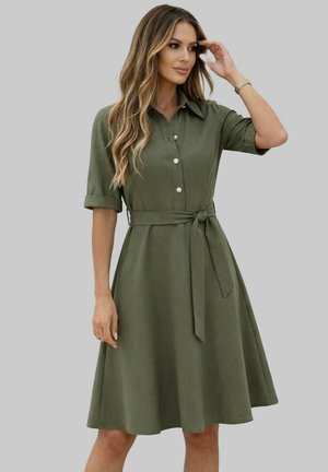 Femme portant une robe-chemise olive longueur genou avec manches courtes et ceinture à la taille, debout devant un fond gris uni.