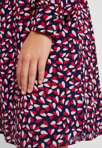 Marinblå klänning med mönster av röda och rosa hjärtformer. Långa ärmar med knäppta manschetter, med en slät tygtextur.