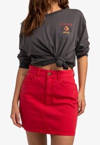 Jupe en denim rouge avec poches avant et un design de couture incurvée, associée à une chemise à manches longues gris nouée avec un imprimé graphique.