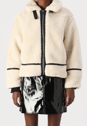 Personne portant une veste crème duveteuse zippée avec un liseré noir et une mini-jupe noire brillante en vinyle sur fond uni.