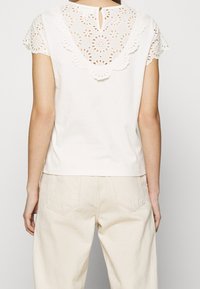 Biały bawełniany top z haftowanymi koronkowymi detalami na ramionach i szyi, z zapięciem na guziki z tyłu. W połączeniu z beżowymi spodniami.