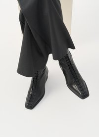 Črne usnjene gležnjarje s koničastim nosom, ki imajo prepleteno vezalno zasnovo in eleganten, gladek videz. Nosi se jih z temno, ohlapno tkanino.