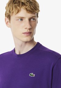 Camiseta de algodón morada con cuello redondo y un pequeño logotipo de cocodrilo verde en la parte izquierda del pecho. Textura suave.