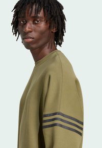 Felpa verde oliva con collo rotondo, maniche ampie e strisce orizzontali nere sulla spalla sinistra. Tessuto di cotone liscio.
