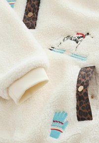 Krémovo sfarbený fleece materiál s výšivkou čierno-bieleho psa, modrým motívom rukavice a leopardím vzorom na zapínaní na gombíky.