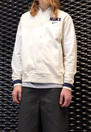 Personne aux longs cheveux portant une veste blanche Nike boutonnée avec un liseré bleu marine et un pantalon noir, se tenant contre un mur noir texturé.
