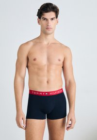 Schwarze Boxer-Briefs mit rotem elastischen Bund, der das Branding "TOMMY HILFIGER" trägt. Glattes Material und körpernahe Passform. Modell ist oberkörperfrei.