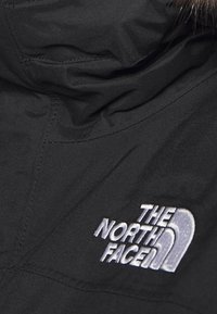 Veste en tissu noir avec capuche et un logo gris brodé bien en vue sur la poitrine, affichant le texte "THE NORTH FACE".