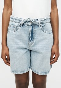Person iført lyseblå denimshorts med selvbindsbælte og synlige forlommer mod en ensfarvet baggrund.