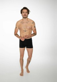 Čierne boxerky na torzálnom mužskom modeli, ktorý predvádza tetovania na rukách a hrudi. Pozadie je jednoduché biele.