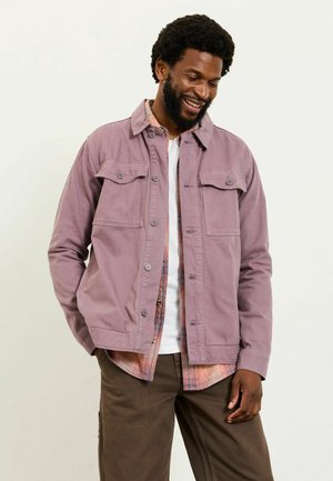 Lila Baumwolljacke mit zwei Vorderentaschen, Knopfverschluss und Kragen. Getragen über einem karierten Hemd in Schattierungen von Orange und Creme.