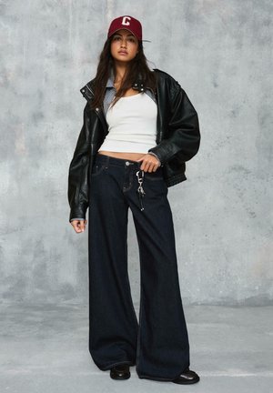 Fiatal nő bordó sapkában, fekete bőrdzsekiben, fehér trikóval, bő szárú sötét farmerral és fekete cipőben, egy szürke falnak támaszkodva.