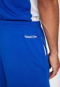 Vista del lado derecho de una persona que lleva puestos unos pantalones cortos azules de Mitchell & Ness y una camiseta deportiva de malla azul y blanca, con la mano relajada a su lado.