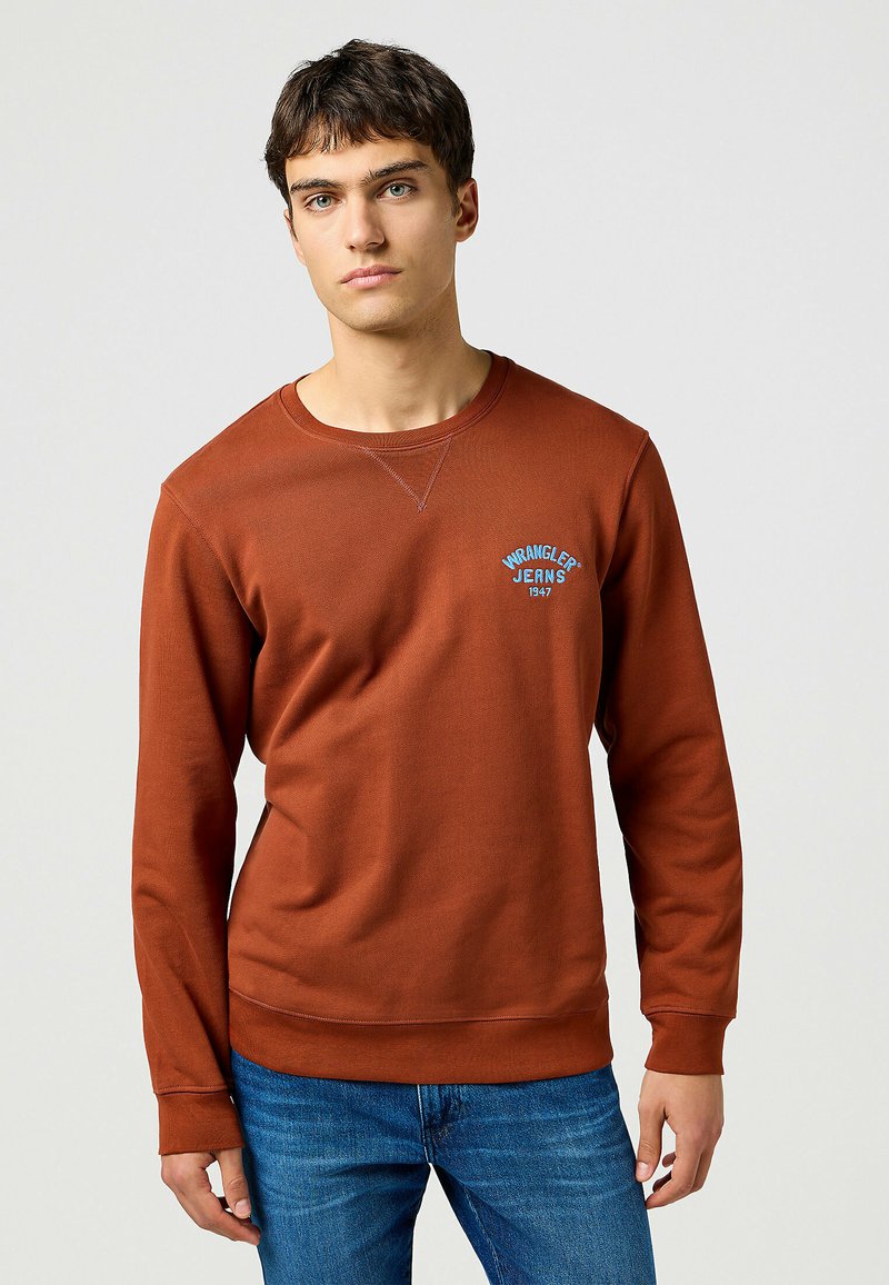 Sudadera marrón de manga larga, cuello redondo, con un logo bordado en azul que dice "Wrangler Jeans 1947" en el pecho. Combinada con unos vaqueros azules.