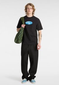 Černé tričko s modro-bílým logem "VANS", spárované s černými kalhotami širokého střihu a zelenou kostkovanou taškou přes rameno. Světle modré boty.