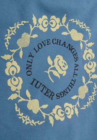 Tessuto blu con ricami floreali dorati che formano un motivo circolare. Testo in nero che recita "SOLTANTO L'AMORE CAMBIA TUTTE LE COSE" e "IUTER."
