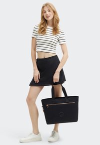 Donna in una gonna nera e un top a righe corto, che tiene una grande borsa tote nera, indossando scarpe da ginnastica bianche, in piedi contro uno sfondo semplice.