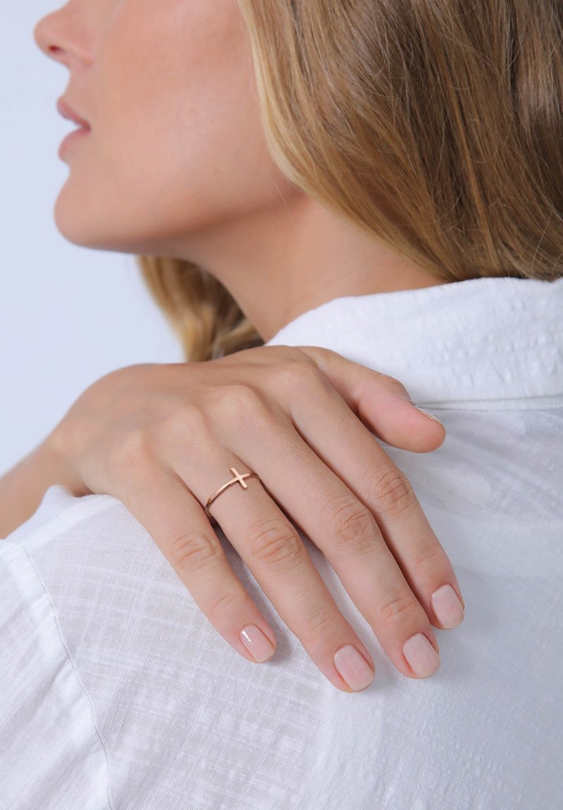 Rosa-goldener Ring mit einem Kreuzdesign auf einer Hand, die auf einem weißen Hemd mit Kragen ruht, mit kurzen, nude-farbenen Nagellack auf den Fingern.