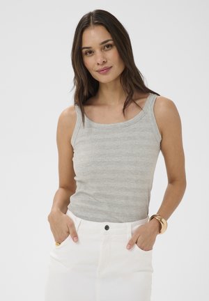 Młoda kobieta o długich ciemnych włosach, ubrana w szary bezrękawnik, białe spodnie, złotą bransoletkę i złoty pierścionek, ręce w kieszeniach, lekko się uśmiecha.