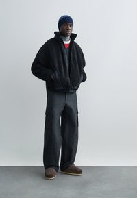 Black fleece jacket with high collar, paired with loose-fitting, dark cargo pants featuring side pockets. Brown patterned slippers complete the look.